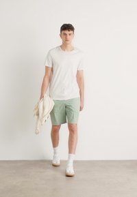 Hvid t-shirt, mintgrønne shorts og hvide sneakers. En lys cremefarvet sweater er lagt over den ene arm. Helkropsvisning mod en neutral baggrund.