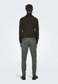 Donkergroene coltrui, aansluitend ontwerp, gecombineerd met grijze broek met zakken en een gestructureerde stof. Zwarte schoenen maken de look compleet.