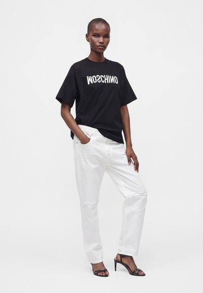 Γυναίκα που φοράει μαύρο t-shirt Moschino, λευκά τζιν με καρδιές στα γόνατα και μαύρα σανδάλια με τακούνι, στέκεται με το ένα χέρι στην τσέπη.