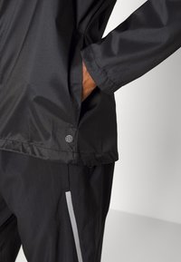 Person som bär en svart jacka med handen i sidofickan och svarta byxor med en vertikal reflektionsrand på låret.