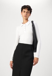 Biely polo top s dlhým rukávom s rebrovanou textúrou a detailom na gombík, v kombinácii s čiernou sukňou s vysokým pásom, ktorá má bočné vrecká a predný rozparok.