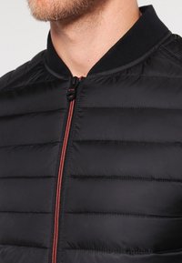 Musta toppatakki, jossa on vetoketju edessä ja vaakasuora tikkaus sekä kontrastivärinen punainen vetoketju. Resorikaulus lisää tekstuuria.