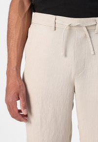 Pantalons en lin beige avec taille à cordon, poches latérales et finition texturée, montrés de mi-cuisse au niveau du poignet.