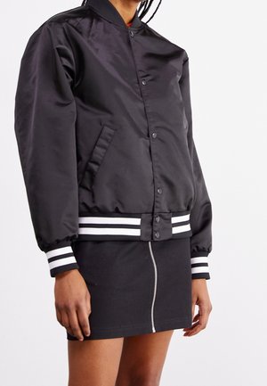 Black satin bomber jacket with a round collar, snap buttons, and white striped ribbing on cuffs and hem. Paired with a black zippered skirt.
