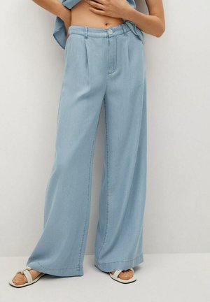 Femme portant un pantalon large bleu clair et des sandales blanches, les mains posées sur le ventre nu, devant un fond uni.