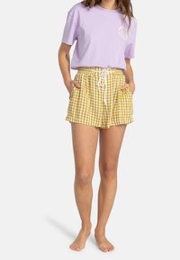 Kvinna i lavendelfärgad t-shirt och gula rutiga shorts med snörning, med händerna i fickorna, stående barfota på vit bakgrund.