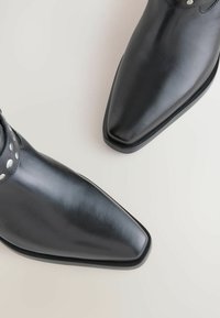 Botas de cuero negras hasta el tobillo con punta afilada, acentos metálicos en la correa, textura suave y suela plana.