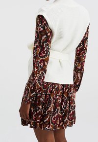 Gilet blanc en maille avec un motif texturé, cintré à la taille, porté sur une robe à motifs floraux aux couleurs bordeaux, verte et crème.