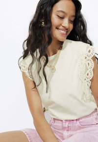 T-shirt en coton beige clair avec un élégant détail en dentelle sur l'épaule gauche, associé à un short en denim rose.