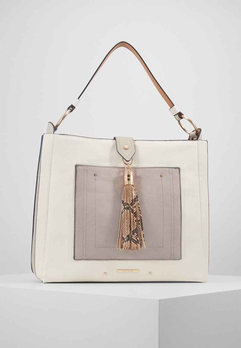 Vit läderhandväska med en beige framficka, guldfärgad metalldetalj och en dekorativ tofs med ormskinnsmönster. Robust handtag i toppen.