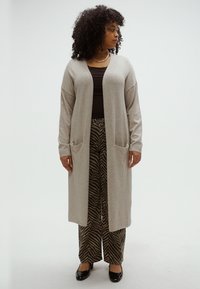 Ανοιχτό γκρι ζακέτα με ανοιχτό μπροστινό μέρος, μακριά μανίκια και δύο μπροστινές τσέπες. Συνδυασμένη με μαύρο και καφέ παντελόνι με σχέδιο και μαύρα παπούτσια.