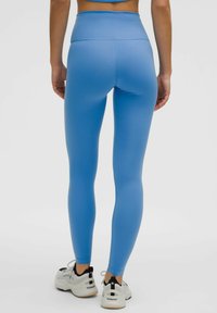 Vysoké modré legíny z elastického materiálu, s hladkým povrchom, priliehavým dizajnom, plochým pásom a detailným prešívaním. V kombinácii s bielymi športovými topánkami.