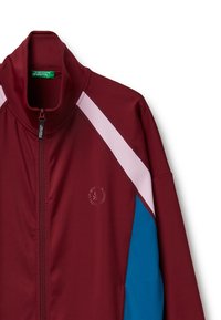 Chaqueta de pista color burdeos con cuello alto, acentos en rosa y azul en los hombros, cierre de cremallera y detalle del logo en el pecho. Fabricada en tejido elástico.