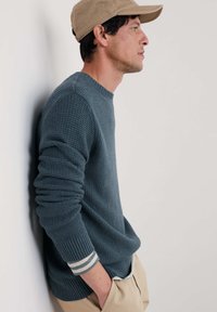 Un pull texture bleu foncé avec des poignets côtelés en rayures grises et blanches, accompagné d'un pantalon clair et d'une casquette beige.