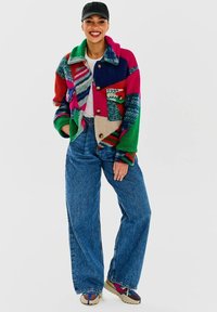 Barevná fleecová bunda s patchwork designem, obsahující červenou, zelenou, modrou a béžovou, v kombinaci s volnými modrými džíny a vzorovanými botami.