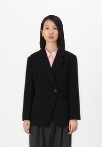 Svart skreddersydd blazer med en knapp, notched lapel og to frontlommer, iført over en lys rosa skjorte og grå bukser.