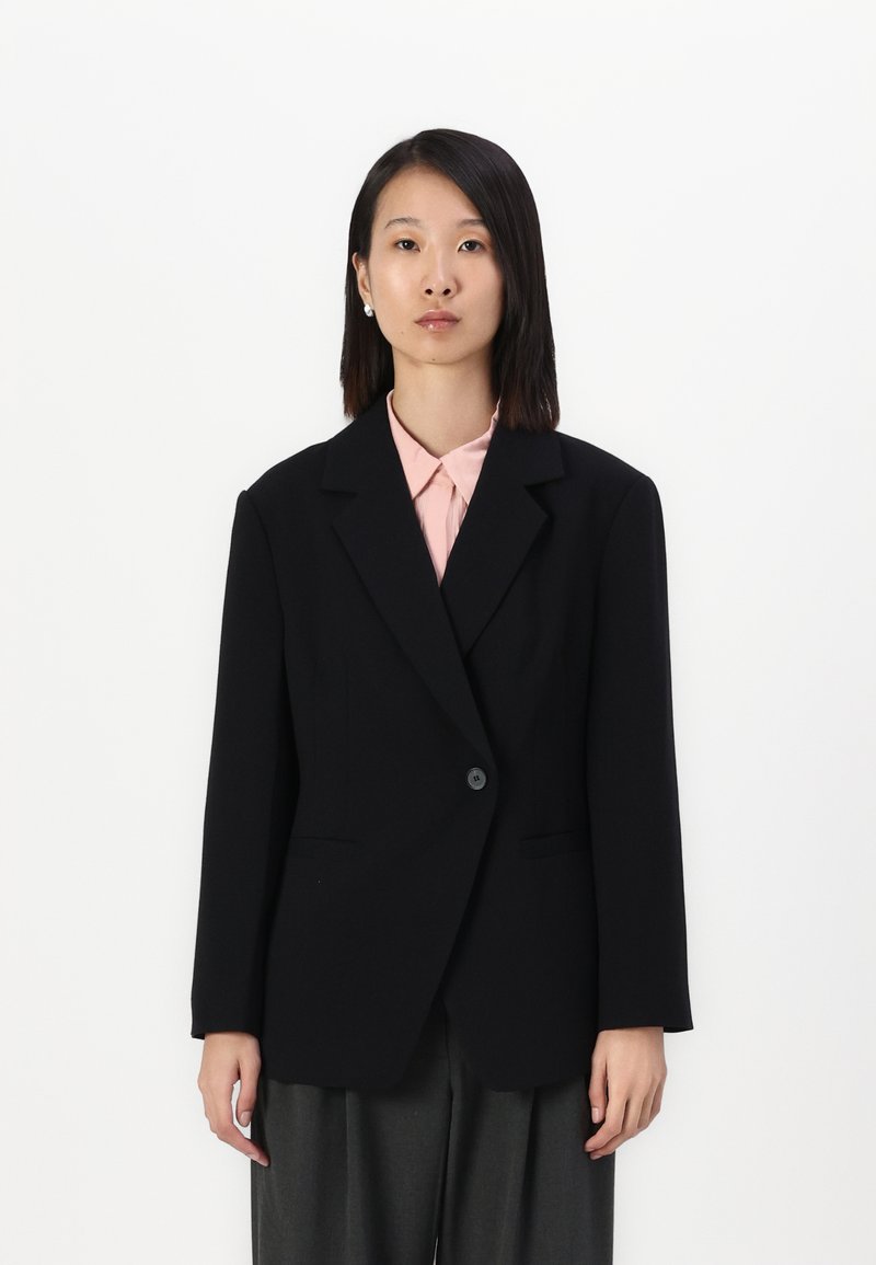 Svart skreddersydd blazer med en knapp, notched lapel og to frontlommer, iført over en lys rosa skjorte og grå bukser.