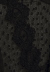 Tissu noir transparent avec un motif texturé de petits carrés. Présente une bordure en dentelle décorative avec des formes en losange et des détails complexes.