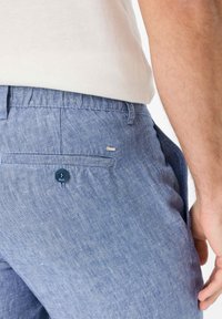 Pantaloni corti in lino azzurro chiaro con una tasca posteriore dotata di un bottone blu scuro e un logo del marchio discreto. La trama è liscia e il fit è casual.