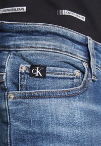 Blå denimjeans med en liten svart etikett med vitt "cK"-logotyp på bakfickan. Sömnadsdetaljer och en urtvättad textur är synliga.