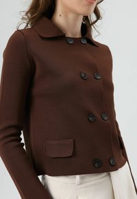 Bruine gebreide jas met kraag, voorzien van zwarte knopen, een dubbelrijig ontwerp en een klein voorvak aan de linkerkant.