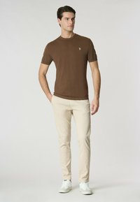 Maglietta corta marrone con colletto rotondo, abbinata a pantaloni slim-fit beige e sneakers bianche. La maglietta presenta un piccolo logo.