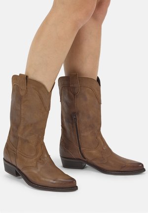 Ben iført brune læder cowboystøvler til midt på læggen med sidelommer og dekorative syninger, vist mod en ensfarvet hvid baggrund.