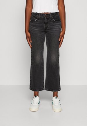 Personne portant un jean noir large raccourci, un crop top blanc et des baskets blanches Adidas avec des bandes vertes, debout devant un fond uni.