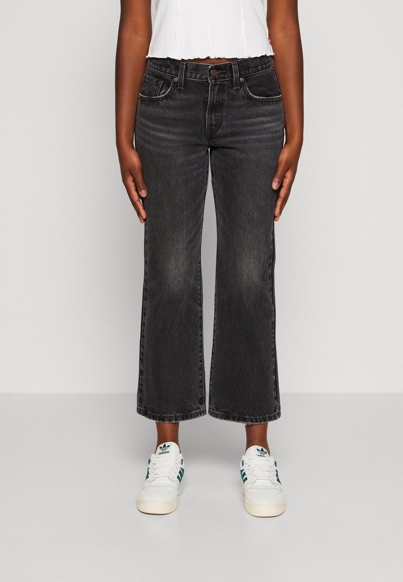 Personne portant un jean noir large raccourci, un crop top blanc et des baskets blanches Adidas avec des bandes vertes, debout devant un fond uni.