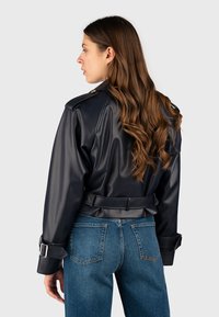 Giacca in ecopelle nera con vita aderente, dettaglio con cintura e spalline, abbinata a jeans blu a vita alta.