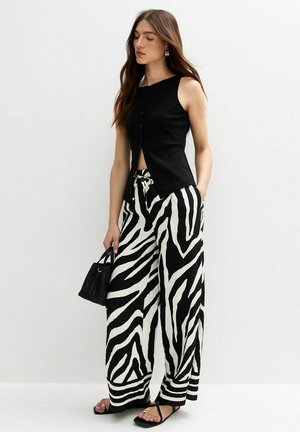 Zwarte mouwloze top met knopen aan de voorkant, gecombineerd met brede zwart-witte zebra gestreepte broek; model houdt een kleine zwarte tas vast.