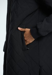 Manteau noir matelassé avec une poche latérale, une surface texturée et des poignets côtelés. Le tissu est lisse et durable.