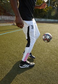 Weiße Sporthosen mit schwarzen Seitenpaneelen und rosa Streifen, kombiniert mit schwarzen Fußballschuhen. Der Spieler dribbelt einen Fußball auf Rasen.