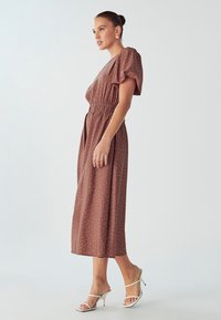 Robe à pois marron avec des manches courtes bouffantes, une taille élastique et une jupe légère. Associée à des sandales à talons blancs. Texture de tissu lisse.