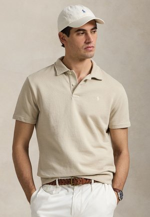Άνδρας που φοράει μπεζ πόλο μπλουζάκι, λευκό καπέλο, λευκό παντελόνι με καφέ πλεκτή ζώνη και ρολόι χειρός, στέκεται με τα χέρια στις τσέπες, κοιτάζοντας προς τα δεξιά.