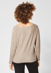 Un haut beige à manches longues avec un large décolleté, une texture douce et une coupe ample, associé à un pantalon noir moulant, vu de dos.