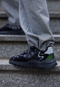 Zapatillas negras con detalles verdes, suela negra texturizada, detalles grises reflectantes y un cordón intrincado, usadas con pantalones de cuadros grises en unas escaleras.