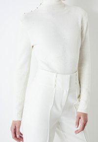 Suéter blanco de cuello alto y mangas largas con un acabado texturizado, adornado con pequeñas cuentas decorativas en el hombro, combinado con pantalones blancos de cintura alta.