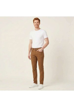 Hombre con pelo rojo y barba, vistiendo una camiseta blanca, pantalones marrones y zapatillas blancas, de pie con una mano en el bolsillo.