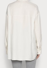Helles beige Langarmhemd mit glatter Textur, ausgestattet mit einem klassischen Kragen und Knopfmanschetten, leicht länger am Rücken.
