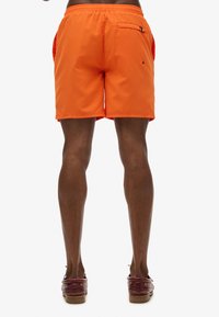Shorts de bain orange avec une taille élastique, poches latérales et une poche arrière zippée. Texture lisse, longueur jusqu’au milieu de la cuisse.