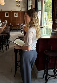 Una persona está de pie en un restaurante sosteniendo un menú, vistiendo una camisa ajustada gris claro y pantalones de pierna ancha gris oscuro. Se pueden ver detalles en madera.