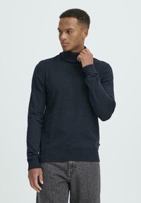 Mørkeblå turtleneck genser laget av myk, ribbestrikket stoff. Har lange ermer og en slank passform. Brukt sammen med grå denimbukser.