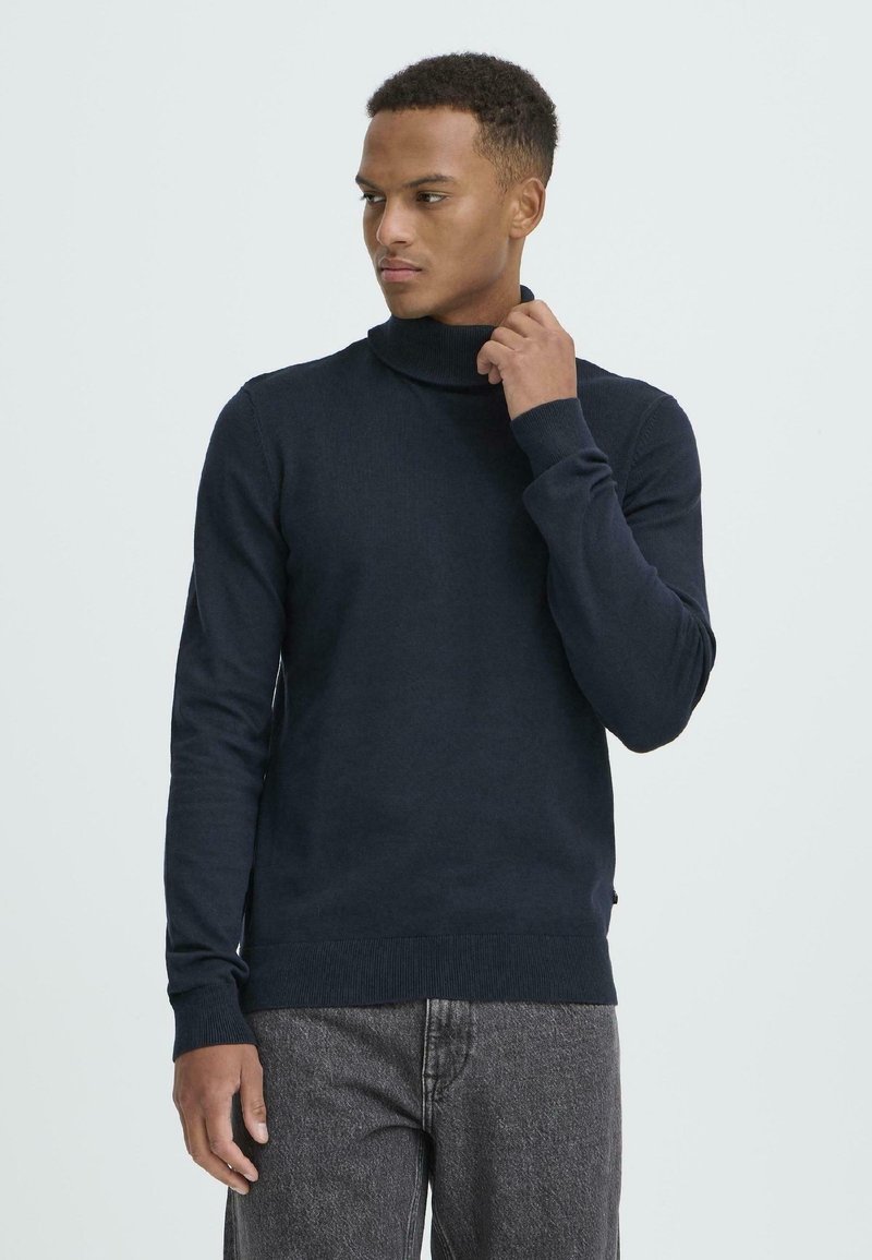 Navy blue turtleneck sweater made of soft, ribbed knit fabric. Features long sleeves and a slim fit. Worn with gray denim trousers.