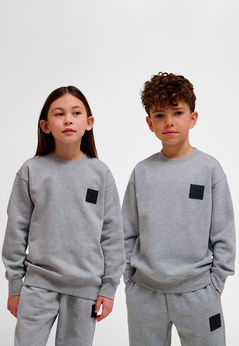 Grå sweatshirt och sweatpants-set; tillverkat av mjukt tyg med en texturerad svart patch på bröstet och matchande byxor. Enkel design med ribbade muddar.