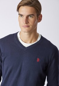 Maglione blu navy a scollo a V realizzato in un tessuto morbido con una sottile trama, presenta un emblema rosso sul petto ed è indossato sopra una camicia bianca.
