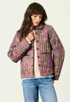 Kvinde med langt brunt hår iført en blomsterquiltet jakke i lilla og pink nuancer, cremefarvet sweater og blå jeans mod en ensfarvet baggrund.