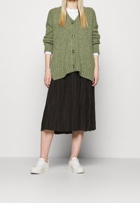 Donna con cardigan spesso verde oliva, gonna midi plissettata nera e sneakers bianche, in piedi su uno sfondo bianco semplice.