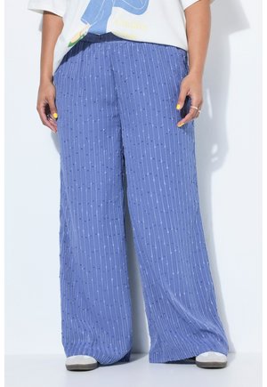 Person trägt lockere, blau strukturierte, weit geschnittene Hosen und weiße Turnschuhe, die Hände entspannt an den Seiten, mit gelbem Nagellack.