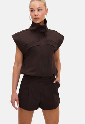 Femme aux cheveux blonds portant un haut noir sans manches à col montant et un short noir assorti, une main dans la poche, regardant légèrement sur le côté.